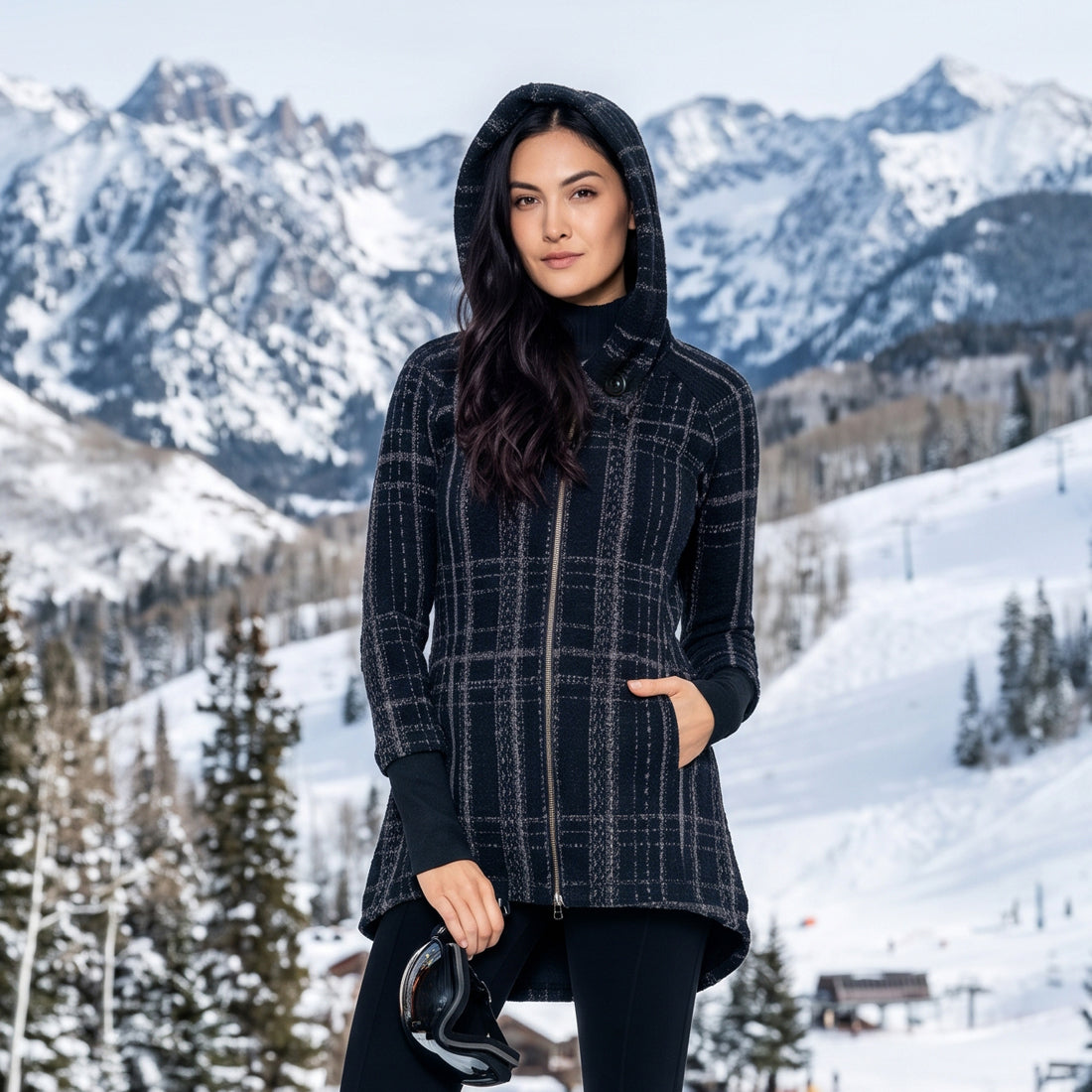 A woman wearing a black hooded jacket with a gray and white plaid pattern stands in a snowy mountain landscape, holding a pair of ski goggles.